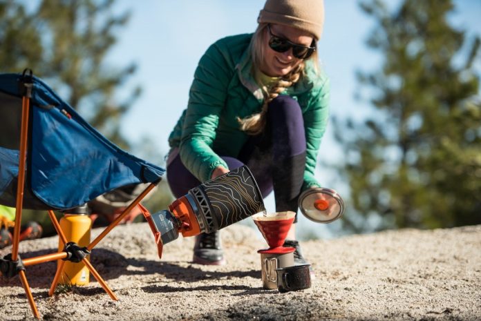 Woman in outdoor setting using a portable camping stove to brew coffee, with a camping chair and mug nearby.